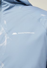 Ανοιχτό μπλε μπουφάν με το λογότυπο Monterrain και εικονίδιο βουνού τυπωμένα με άσπρο χρώμα στο πάνω μέρος της πλάτης.