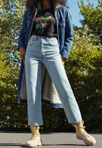 Ljust blå högmidjade jeans kombineras med en svart t-shirt med tryck och en lång jeansjacka. Gula snörstövlar fulländar outfiten.