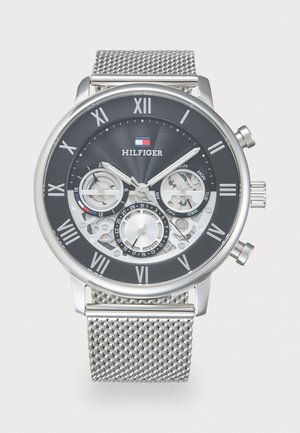 Silver mesh strap watch with a black face, silver Roman numerals, and visible gears. Features include dual time zones and day indicators.