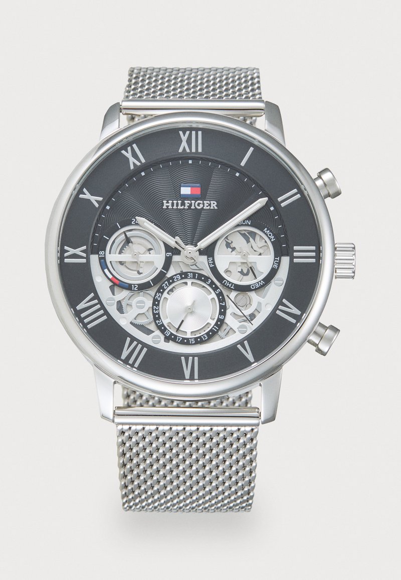 Silver mesh strap watch with a black face, silver Roman numerals, and visible gears. Features include dual time zones and day indicators.