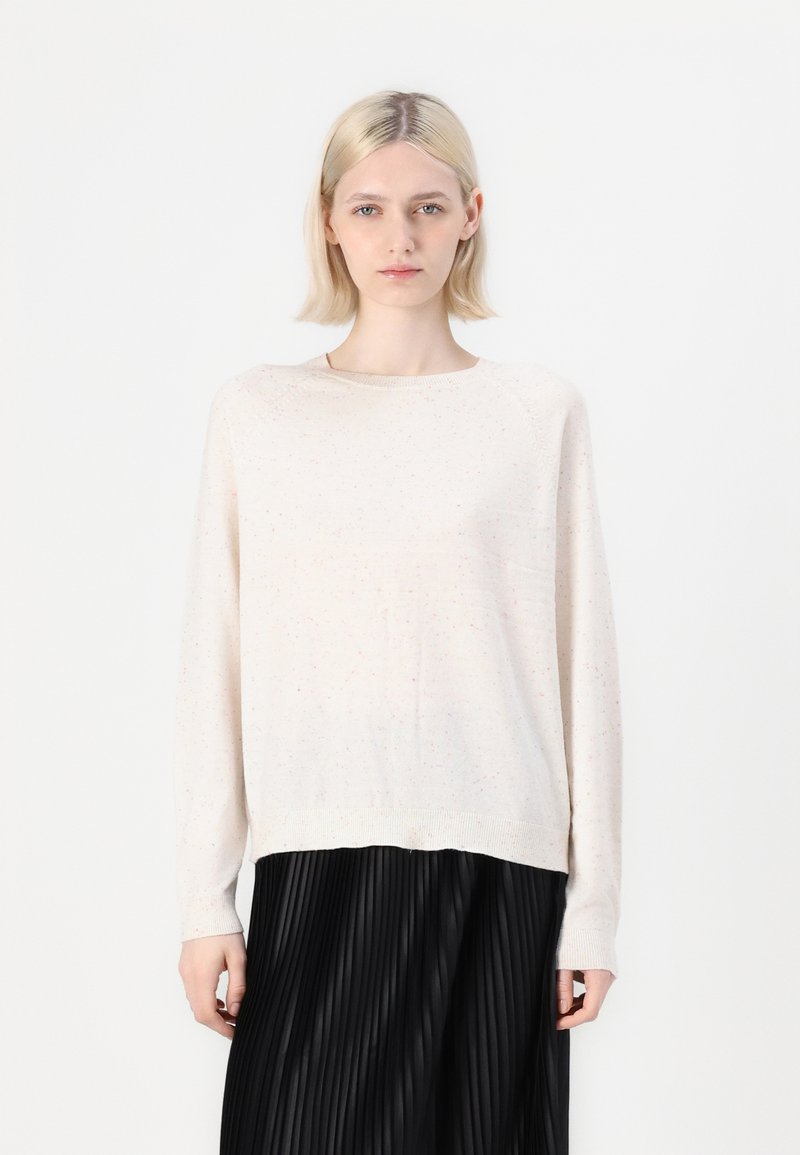 Blonde Frau in einem hell gesprenkelten Pullover und einem schwarzen Faltenrock, die vor einem schlichten weißen Hintergrund steht.