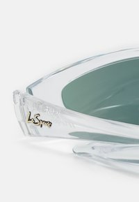Occhiali da sole in plastica trasparente con lenti verdi, caratterizzati da una forma curva e un accento dorato con il logo "Le Specs" sulla tematica.