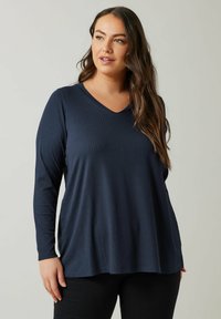 Top a maniche lunghe ribbed blu navy con scollatura a V. Il tessuto ha una texture morbida e una vestibilità rilassata, che si abbina a dei pantaloni neri.