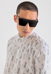 Gafas de sol negras de gran tamaño con montura angular, llevadas por una persona con una camisa de color claro y patrones de pinceladas negras.