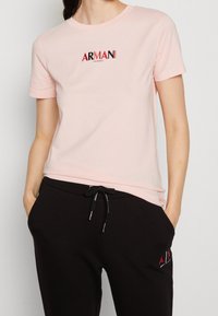 Osoba ubrana w jasnoróżową koszulkę Armani Exchange i czarne spodnie z troczkami, pokazana od szyi do bioder, z rękami w kieszeniach.