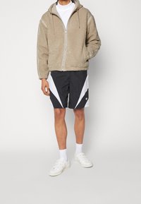 Beige fleece zip-up hoodie med fodrad huva, kombinerad med svarta shorts med vita diagonala detaljer. Vita sneakers och korta strumpor fulländar looken.