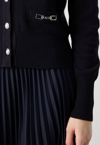 Cardigan tricoté bleu marine avec un ourlet côtelé, des boutons argentés, et une poche ornée d'une chaîne. Tissu plissé visible en dessous.