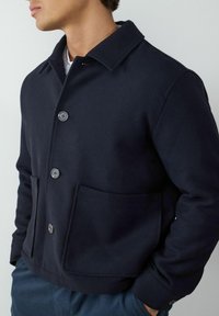 Granatowa kurtka wykonana z miękkiego materiału, z przodem zapinanym na guziki, dużymi kieszeniami i klasycznym kołnierzem. Gładka tekstura, swobodny krój.