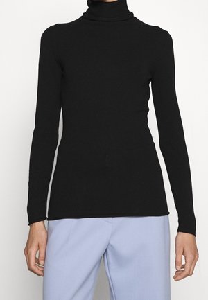 Femme portant un pull noir ajusté à col roulé et manches longues et un pantalon ample bleu clair, debout avec les bras détendus le long du corps.