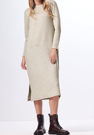 Woman wearing a beige long-sleeve midi sweater dress with side slits and dark brown lace-up ankle boots, standing against a white background.