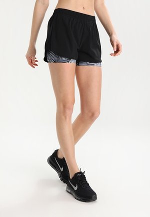 Mujer con pantalones cortos deportivos negros en capas y zapatillas negras de running Nike, de pie frente a un fondo blanco liso.