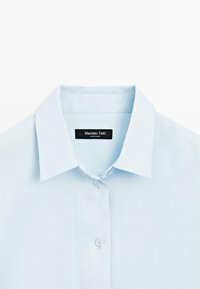 Camicia azzurra chiara con bottoni, realizzata in tessuto liscio. Presenta un colletto strutturato, due bottoni frontali e un'etichetta del marchio all'interno del colletto.