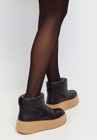Piernas en medias negras transparentes que visten botas de tobillo negras con suelas gruesas beige y forro de leopardo, de pie sobre un suelo blanco.