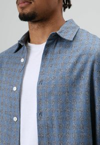 Geruit blauw-grijs overhemd van geweven stof, met een knoopsluiting aan de voorkant, een brede kraag en een contrasterend wit T-shirt eronder.