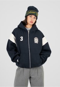 Námornícky sveter s bielymi akcentovými pruhmi, zipsom, vyšitým logom a zipsovou bočnou vreckou. Kombinovaný so šedými plisovanými nohavicami a čiapkou.