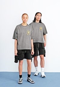 Camisetas gris oversize con un gráfico circular amarillo, combinadas con pantalones cortos negros. El escenario presenta un fondo blanco y un suelo azul.