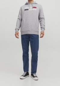 Felpa grigia con tasca frontale e logo testuale, abbinata a jeans di denim blu e sneakers nere con suole bianche. Design casual.