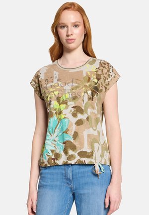 Junge Frau trägt ein beiges T-Shirt mit floralem Muster und dem Schriftzug "Good Vibes" sowie eine blaue Jeans, steht vor einem einfarbigen Hintergrund.