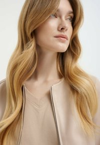 Beige kožená bunda se stříbrným zipem, spárovaná s béžovým topem s výstřihem do V. Dlouhé, vlnité blond vlasy padají přes ramena. Neutrální pozadí.