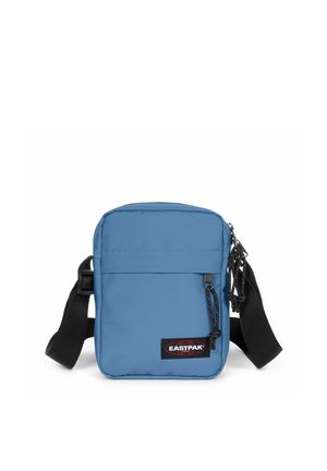 Bolso pequeño azul Eastpak con correa negra ajustable y bolsillo frontal con cremallera sobre fondo blanco.