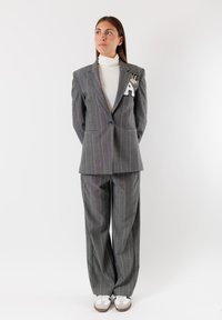 Completo grigio a righe con giacca aderente, caratterizzata da una lettera "A" ricamata e un emblema. Pantaloni a gamba larga coordinati e sneakers bianche.