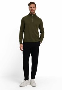 Maglione verde a maglia con colletto alto e zip argentata, abbinato a pantaloni neri e sneaker bianche. Texture liscia e design aderente.