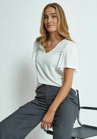 Top bianco a scollo a V con maniche corte a sbuffo, abbinato a pantaloni grigi a vita alta, seduta su una sedia metallica. Tessuto dalla superficie liscia.