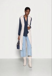 Casaco de trench sem mangas bege sobre uma blusa azul-marinho, combinado com jeans de perna larga azul-claro e botas de salto bege. Bolsa azul na mão.