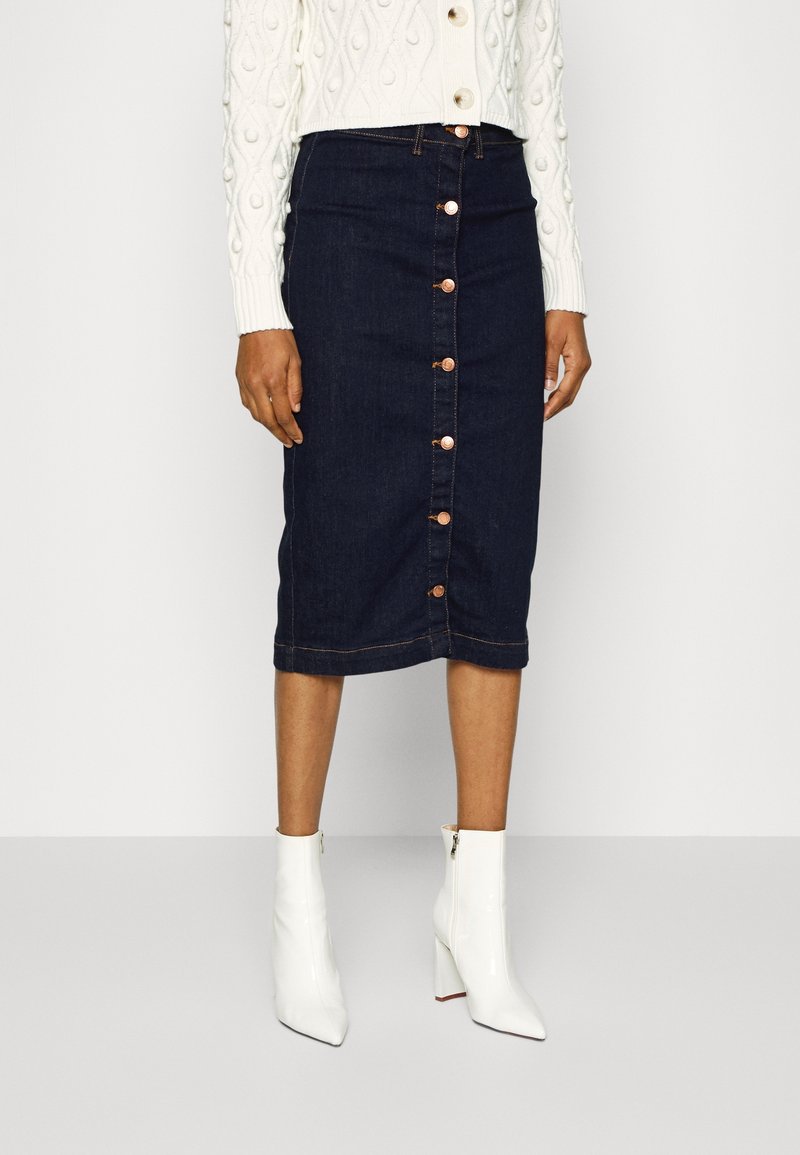 Denim pennkjol i mörkblått med knappstängning fram och kopparknappar. Kombinerad med en vit stickad tröja och vita klackade ankelboots.