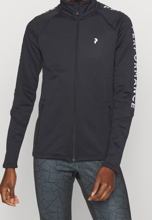 Veste noire zippée avec logo blanc sur la poitrine et texte "PERFORMANCE" sur la manche gauche portée sur un legging foncé à motifs.
