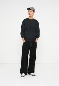 Joven vestido con un suéter de punto oscuro, pantalones negros de gran tamaño, gorra gris y zapatillas blancas, de pie con las manos en los bolsillos.
