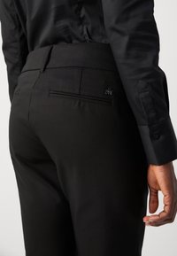 Un pantalon noir sur mesure présente un tissu lisse, une coupe slim et une poche arrière. La ceinture est plate avec des détails de couture subtils.