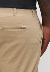 Bež pamučne hlače s stražnjim džepom koji ima bijelu oznaku s natpisom "JACK & JONES." Glatka tekstura i kroj koji odgovara.