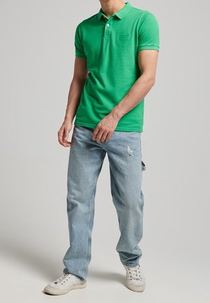 Homme portant un polo à manches courtes vert vif, un jean bleu clair déchiré, et des baskets blanches, debout devant un fond gris uni.