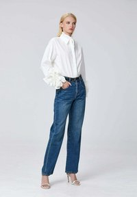Chemise blanche à manches volantées, associée à un jean taille haute bleu et à des sandales à talons hauts blanches à lanières. Arrière-plan simple.