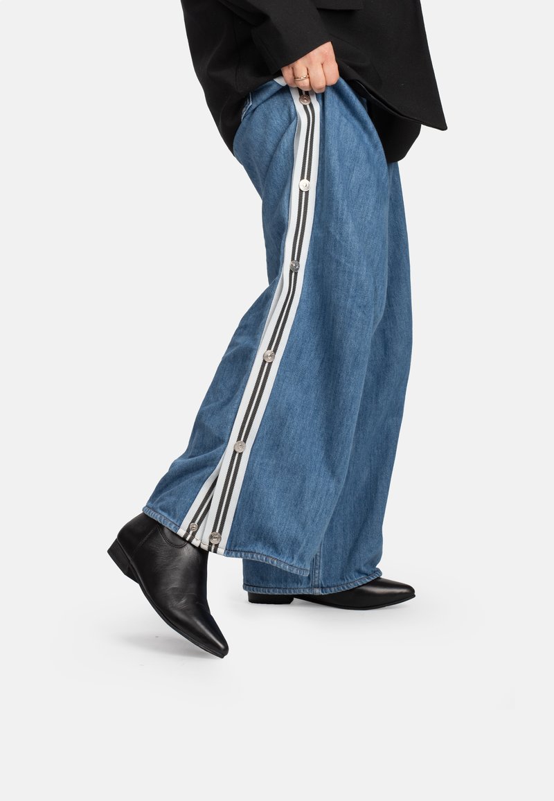 Pantaloni larghi in denim blu con pannelli laterali a strisce bianche e nere, caratterizzati da dettagli con bottoni e indossati con stivaletti neri alla caviglia.