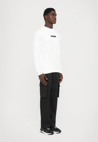 Ragazzo giovane in piedi, vestito con un felpa bianca con logo nero, pantaloni cargo neri con grandi tasche e scarpe sportive bianche e nere.