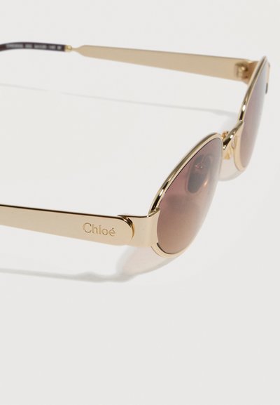 Γυαλιά ηλίου με χρυσό μεταλλικό πλαίσιο και φακούς καφέ απόχρωσης, με την εγχάρακτη επιγραφή "Chloé" στο πλαϊνό μπράτσο, εμφανιζόμενα σε λευκό φόντο.