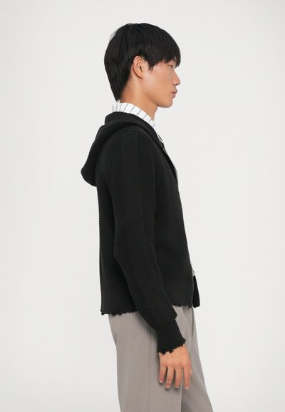 Svart stickad hoodie med ett texturerat mönster och fransad fåll, bärs över en collared skjorta med bl�å ränder, visad från sidan.