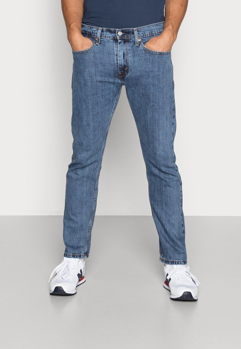 Homme portant un jean en denim bleu et des baskets blanches, debout avec les mains dans les poches devant un fond uni.