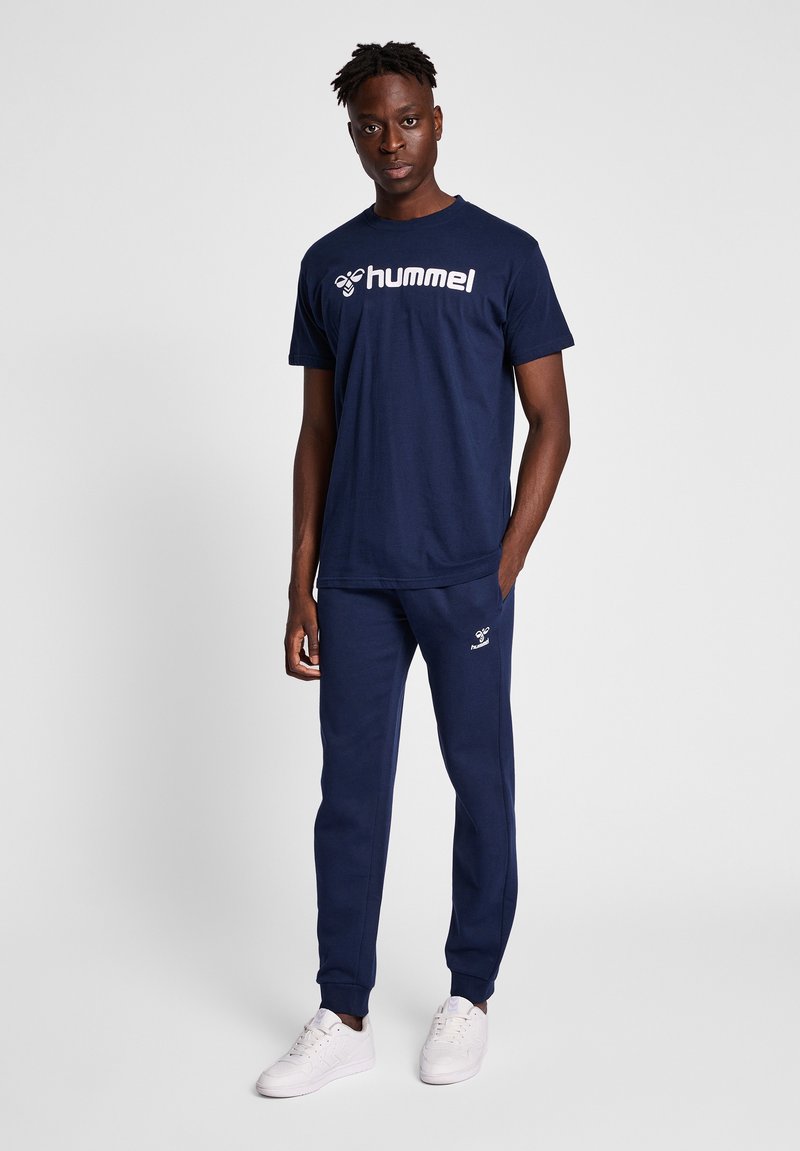 Marineblaue Baumwoll-T-Shirt mit "hummel"-Logo, kombiniert mit passenden marineblauen Sweatpants. Das Model trägt weiße Sneaker. Schlichtes, sportliches Design.