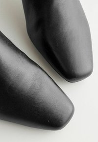 Gros plan sur les bouts de deux chaussures en cuir noir de forme carrée, posées sur une surface claire.