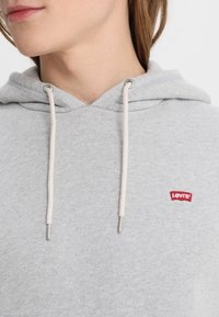 Persona vistiendo una sudadera ligera gris con cordones blancos y un pequeño logo rojo de Levi's en el pecho, mostrando la parte inferior de la cara y el cuello.