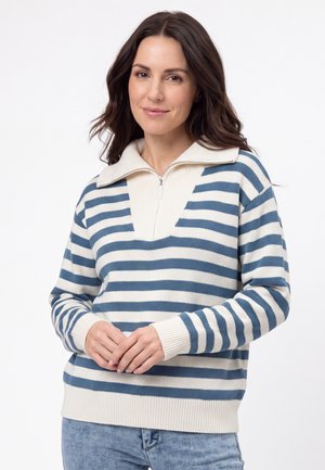 Donna con capelli castani lunghi che indossa un maglione a righe bianche e blu con zip e jeans azzurri chiari, in piedi con le mani conserte.