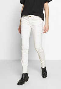 Jeans skinny de denim blanco con detalles desgastados, cierre de botón, cinco bolsillos y un corte ajustado. Combinados con botas negras hasta el tobillo.