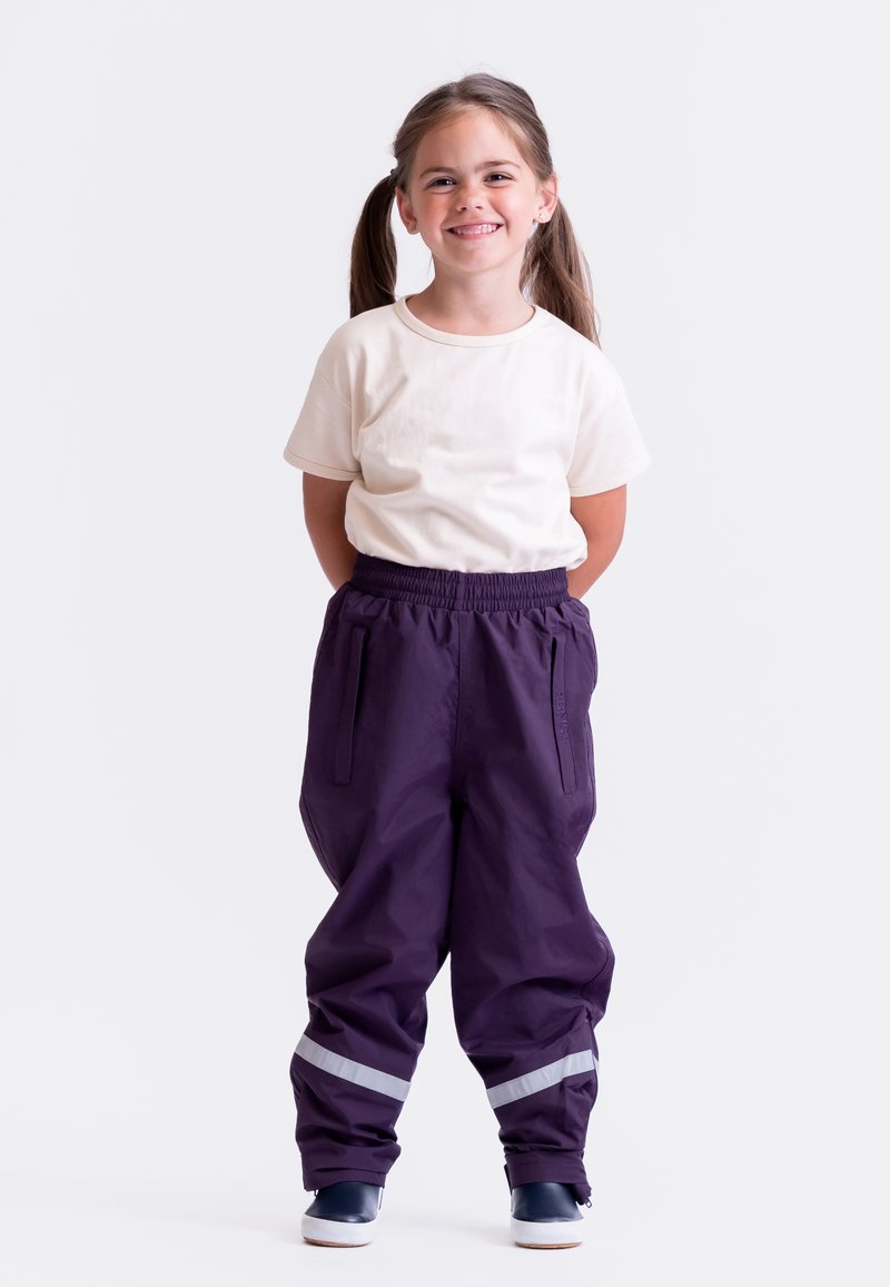 Lila Windpants mit elastischem Bund, reflektierenden Akzenten am Saum, kombiniert mit einem schlichten, hellbeigen T-Shirt und blauen Schuhen.