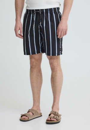 Man draagt zwarte zwembroek met verticale witte en blauwe strepen, beige sandalen en een wit ingestopt overhemd, staand tegen een effen achtergrond.