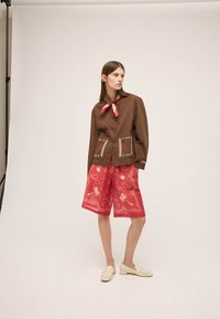Žena stojac v hnedom zapínanom saku s vyšívanými vreckami, červených šortkách po kolená so vzorom, červenom šatkovi na krku a bielych mokasínach na jednoduchom pozadí.
