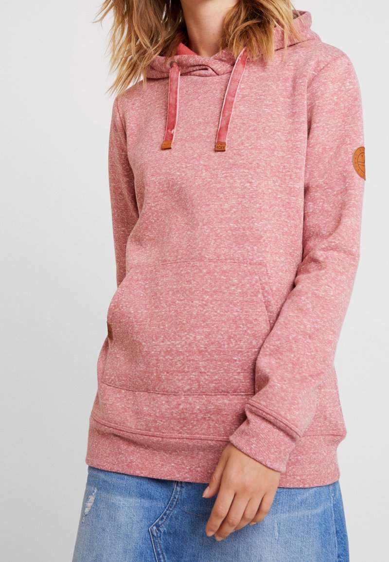 Woman wearing a pink speckled hoodie with front pocket and drawstrings paired with blue denim jeans.