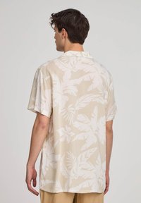 Camicia a maniche corte in tessuto beige chiaro con un motivo di foglie tropicali bianche, caratterizzata da una vestibilità rilassata e colletto.